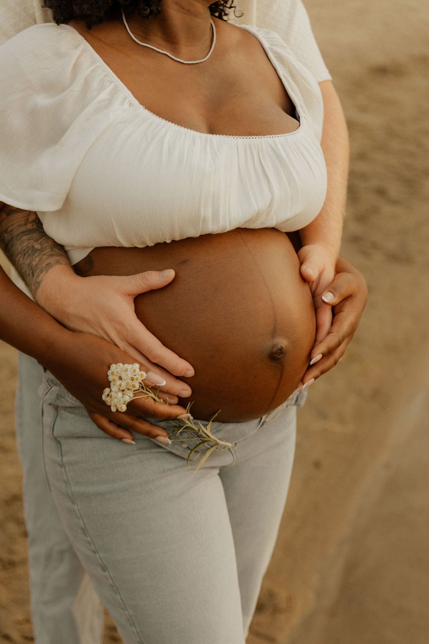 Close-up of pregnant belly being cradled in white blouse, wearing statement floral ring and casual light colored pants.