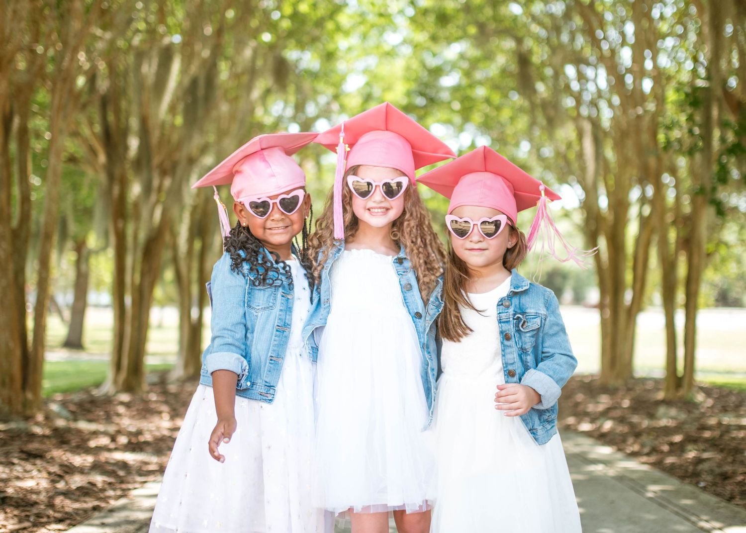 Three preschool girls wearing pink graduation caps, denim jackets, and white dresses at Yellow Jacket Park in Orlando, FL. Cred: LexTakePix