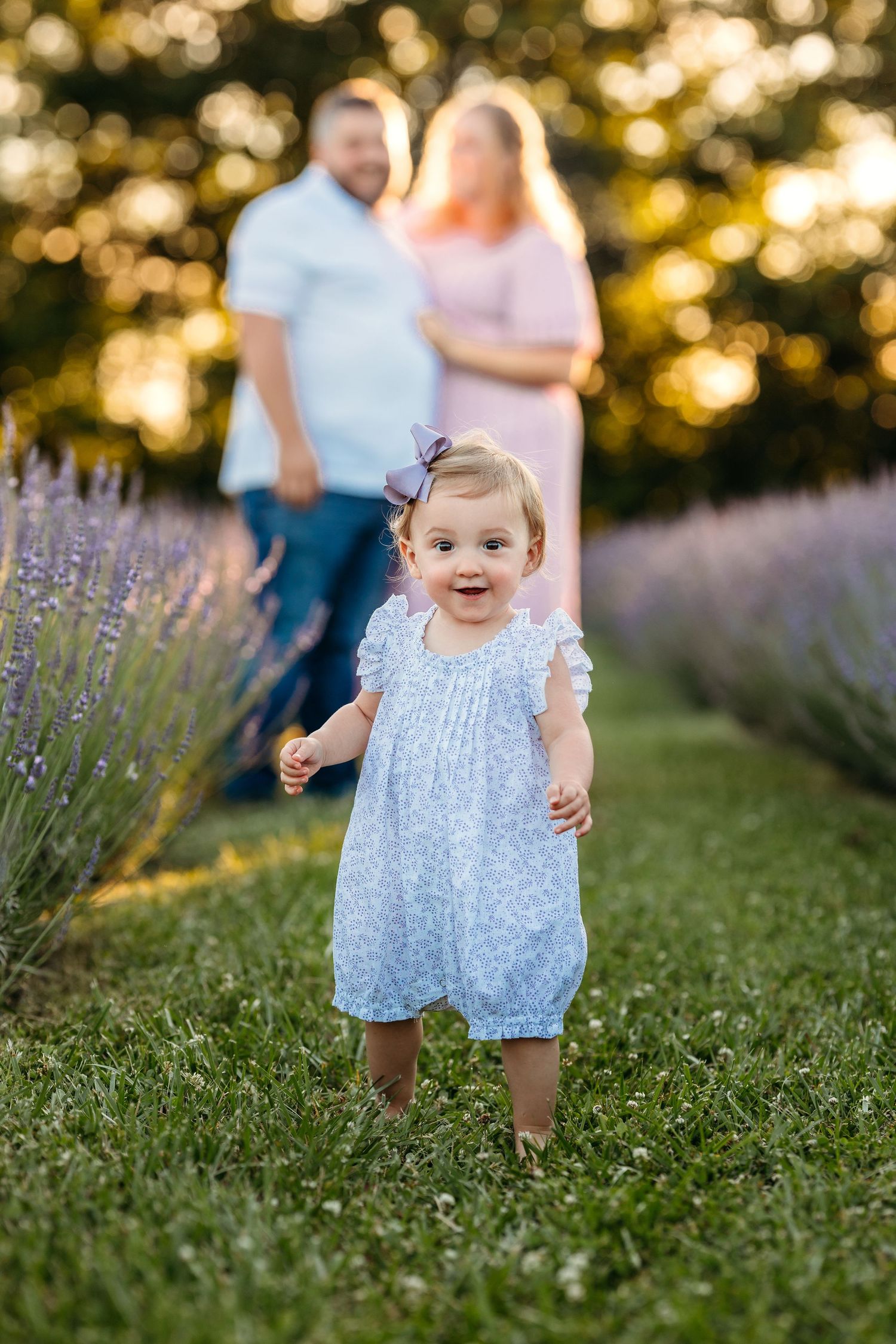 A toddler in a blue floral dress walks through a garden with lavender flowers and bokeh lighting at sunset.  Middle Tennessee Luxury Child P