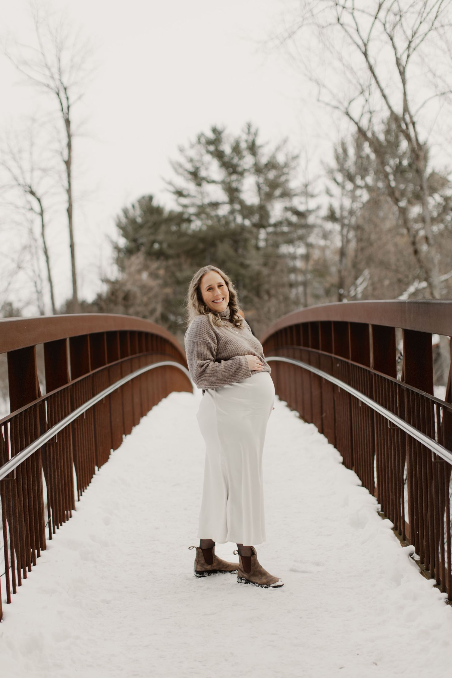 A person in a white dress and brown boots sits on a rustic metal bridge during winter.