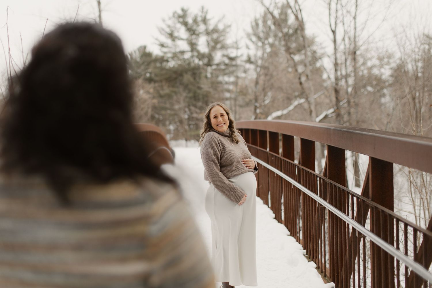 A couple in winter coats holds hands while walking across a rustic wooden bridge in a snowy landscape.