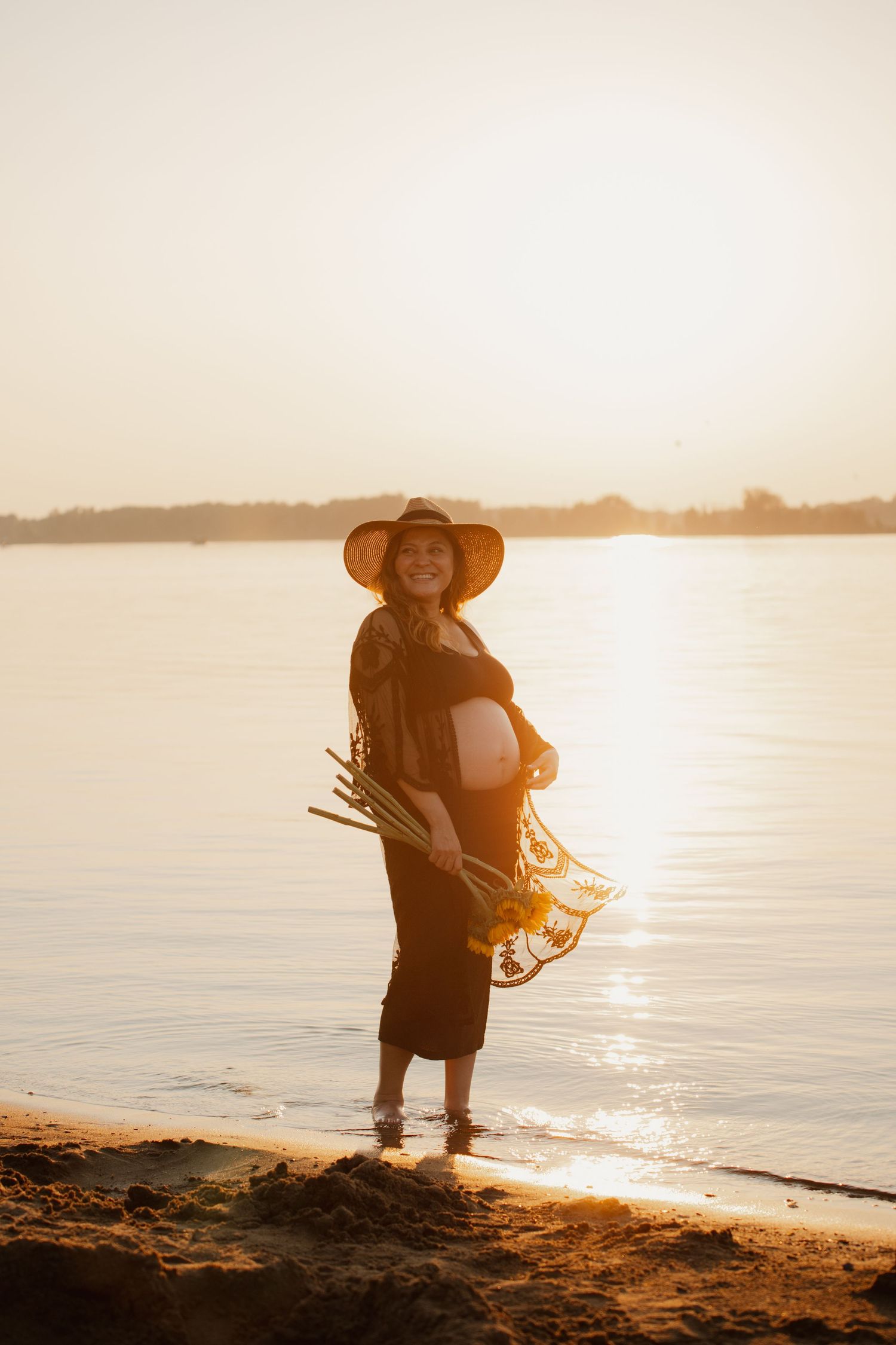Golden hour maternity portrait on beach with sunlit water background.