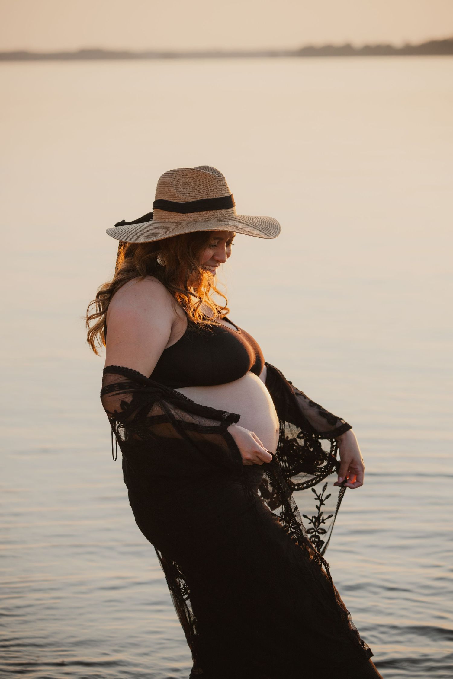 A maternity photoshoot at sunset shows an elegant silhouette in a black lace dress and wide-brimmed hat by the water.