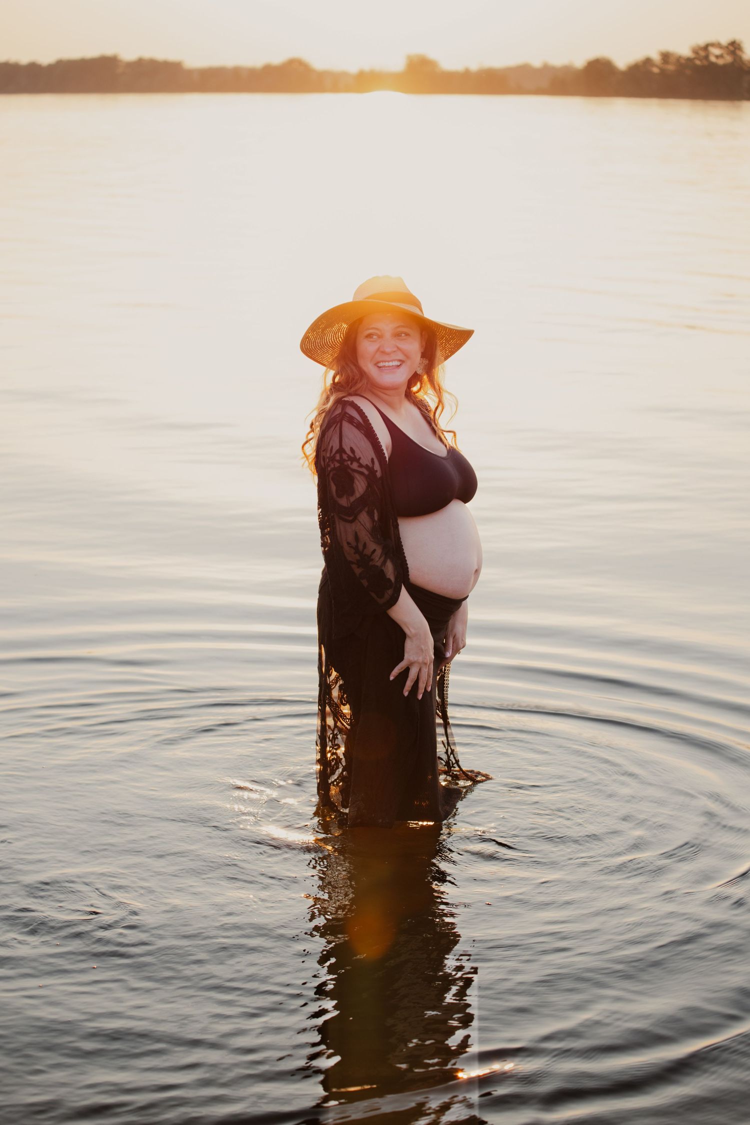 A sunset maternity photo captures a moment standing in calm lake waters wearing a black lace dress and sun hat.