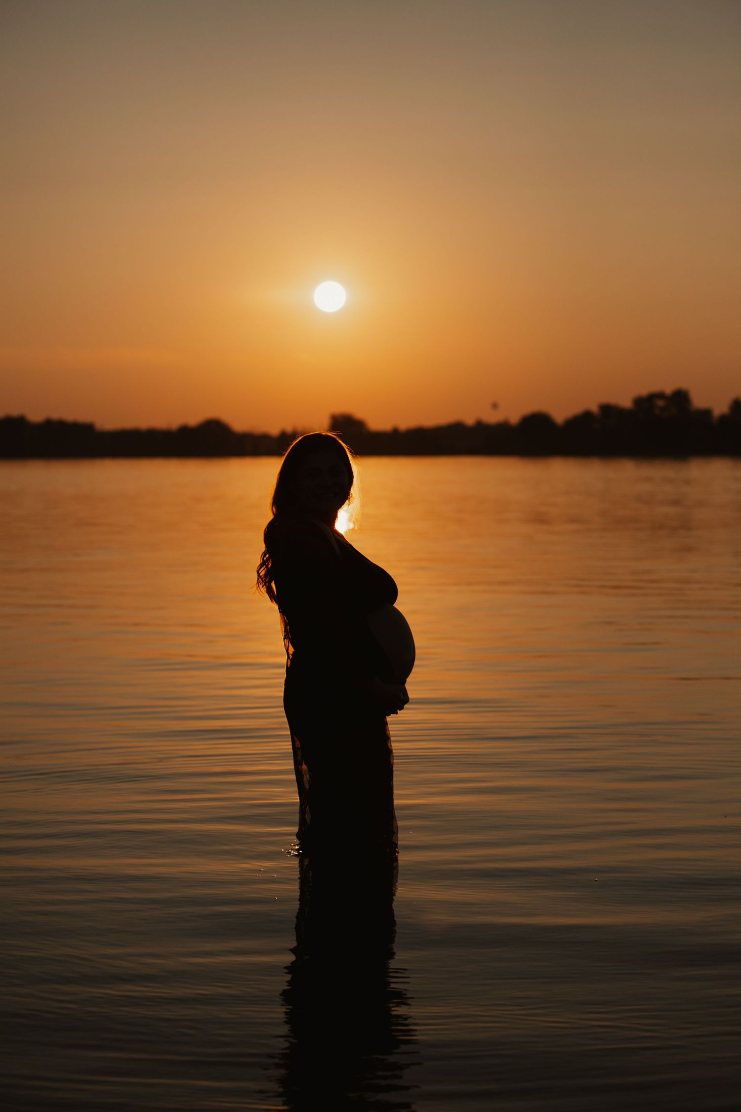 Silhouette of an expectant mother standing in water against a dramatic orange sunset sky.