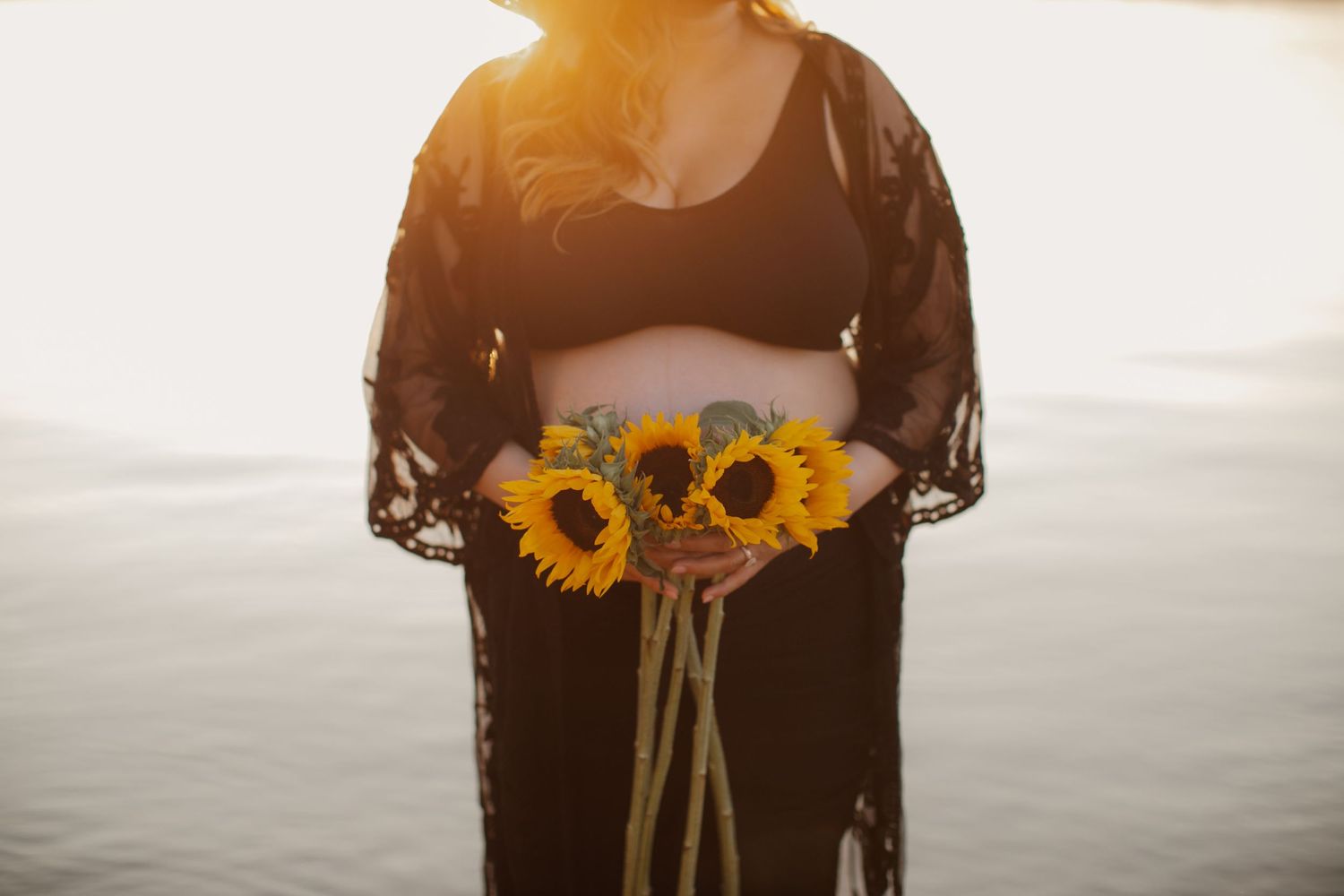 Silhouette of pregnant woman holding sunflowers against sunset water backdrop.