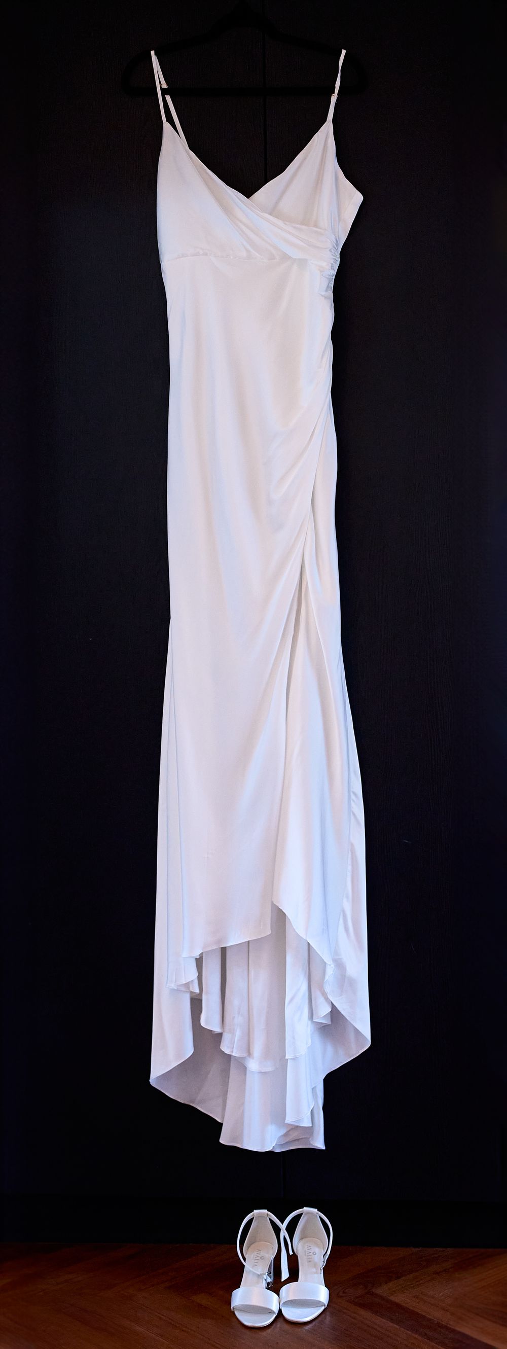 Elegant white wedding dress with spaghetti straps and high-low hem hanging against dark background with matching heels below.