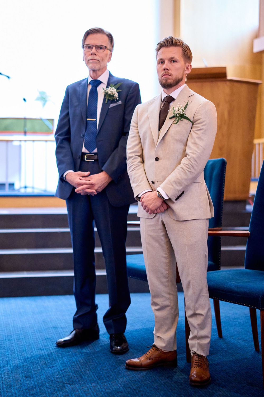 Two men stand together at a wedding ceremony, wearing contrasting suits of navy blue and light beige.