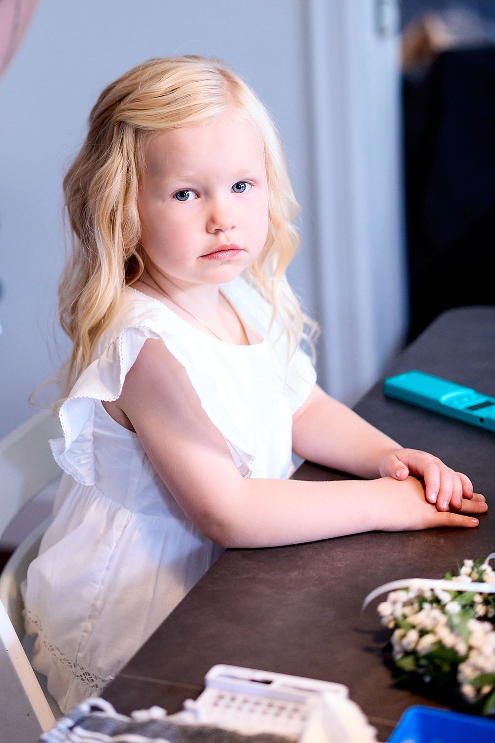 A young child in a white ruffled dress sits at a dark wooden table with flower arrangements nearby.