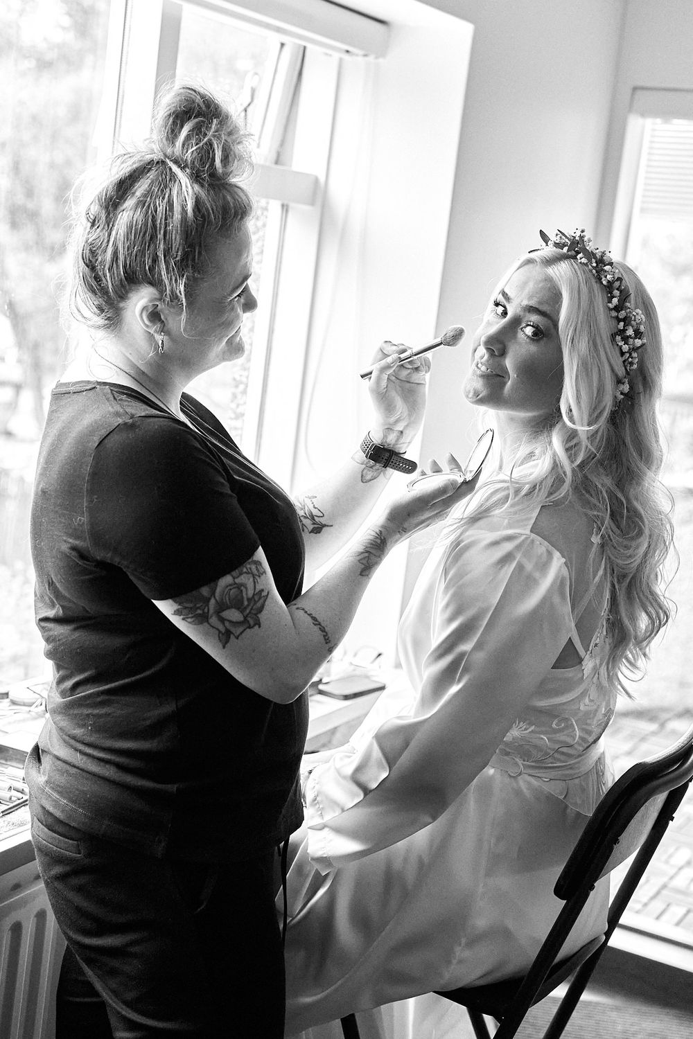 A makeup artist applies cosmetics to a bride wearing a floral crown during wedding preparations.