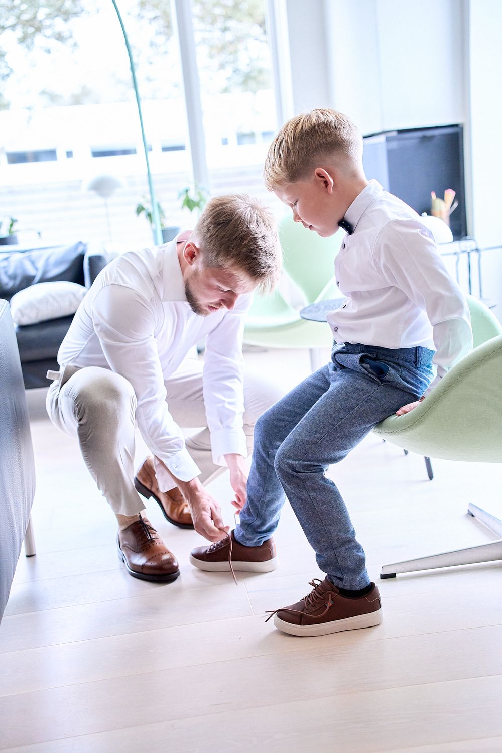 Parent helping child tie shoelaces in bright modern room with white and mint green furniture.