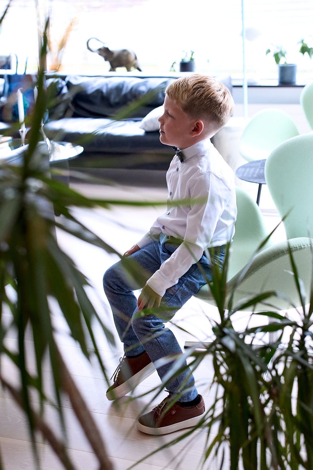 A child in a white shirt and jeans jumps playfully in an indoor setting with plants visible.