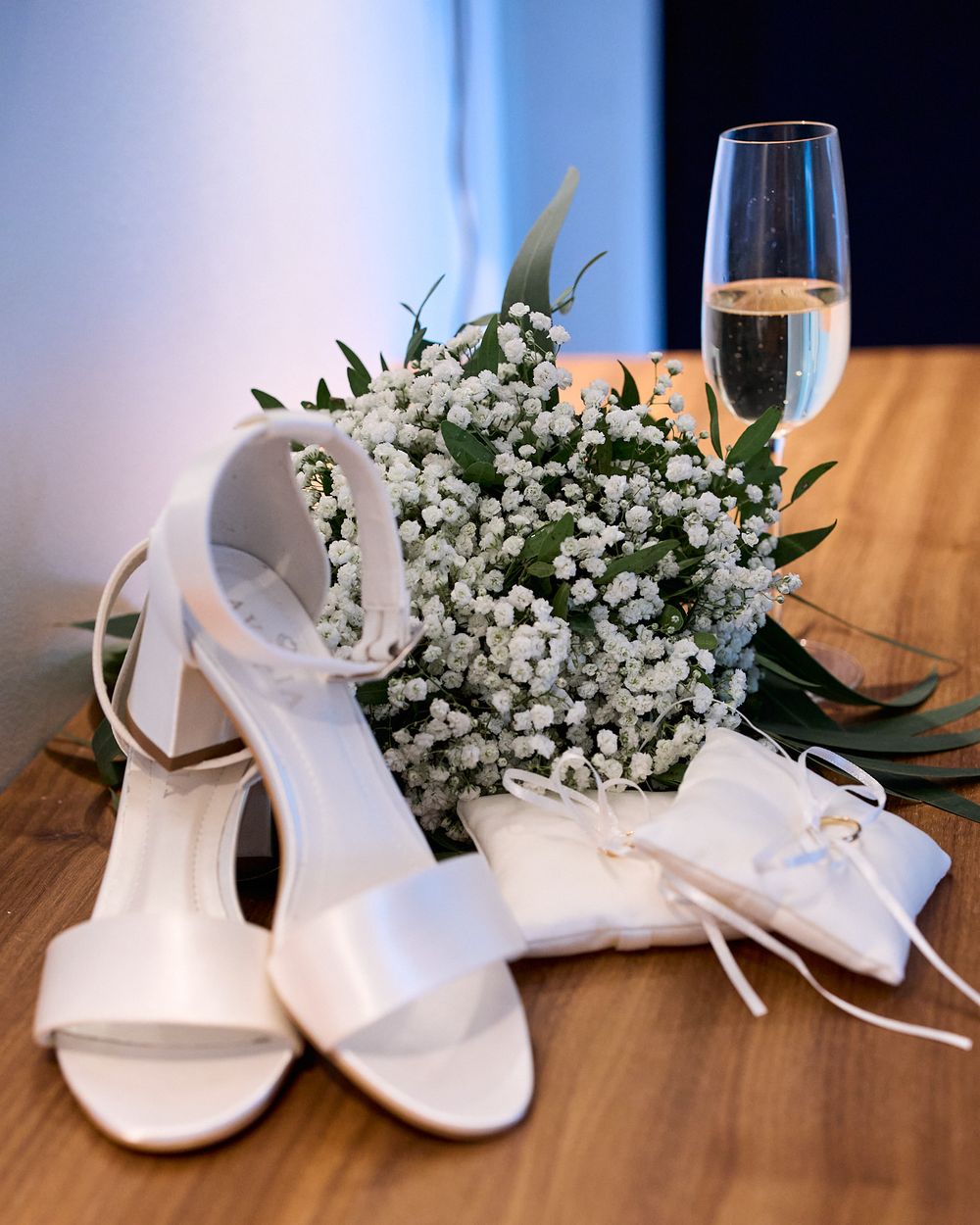 Wedding accessories including white heels, baby's breath bouquet and champagne glass arranged on wooden surface.