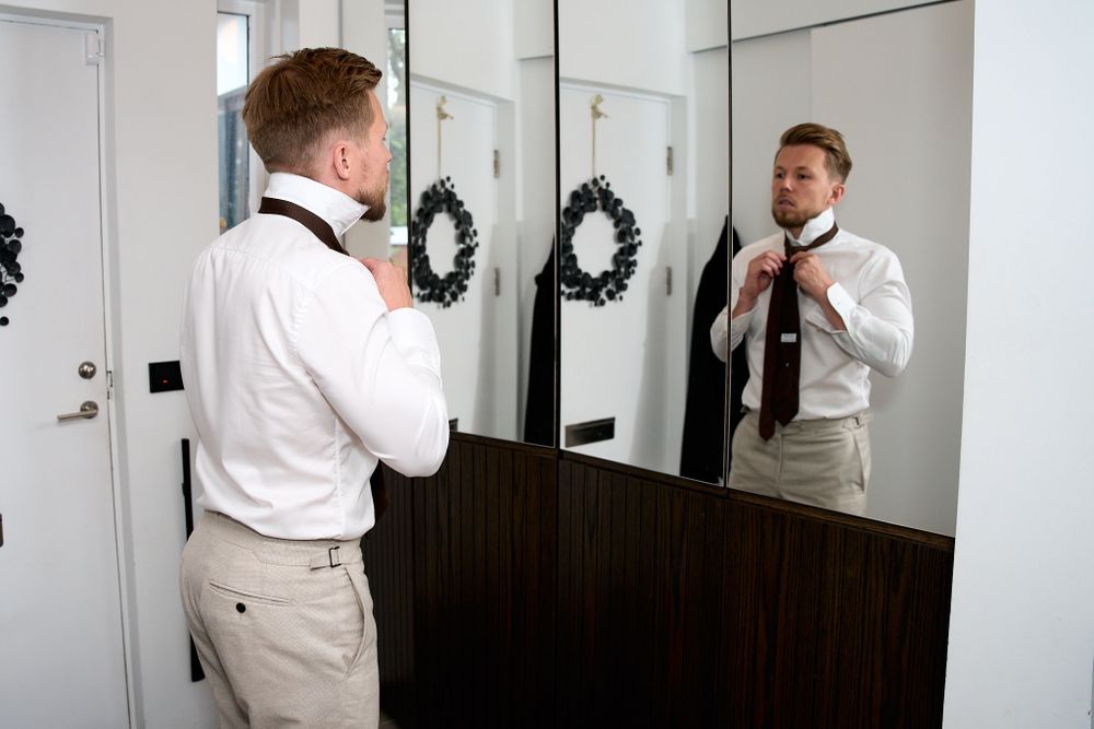A person in formal attire adjusts their tie while looking in a mirror with dark wreaths hanging on nearby doors.