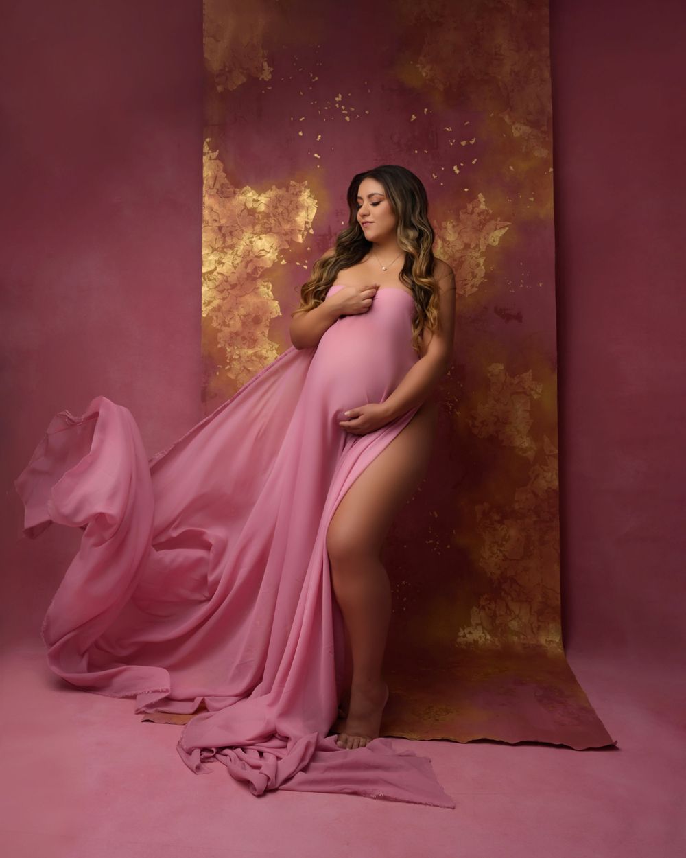 Romantic maternity portrait with flowing pink fabric against textured burgundy and gold backdrop.