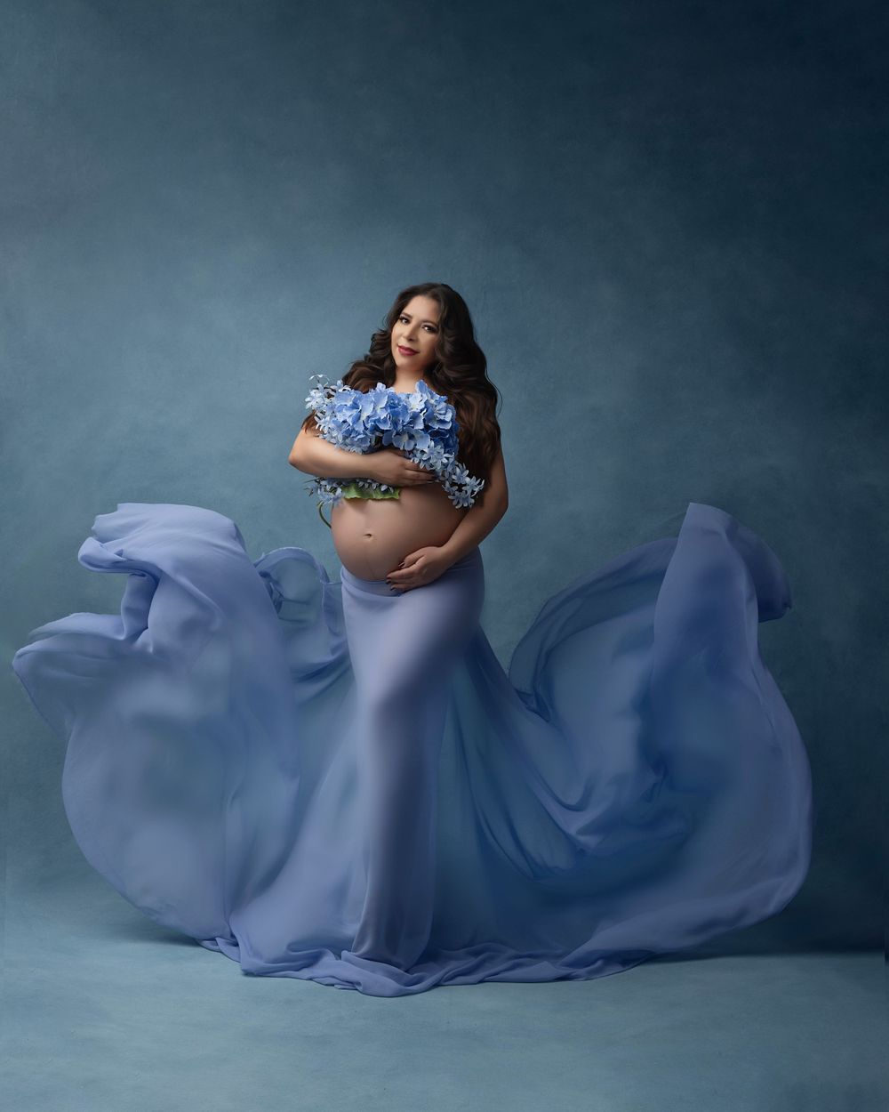 Maternity photoshoot featuring flowing periwinkle fabric creating elegant movement against a moody blue backdrop.
