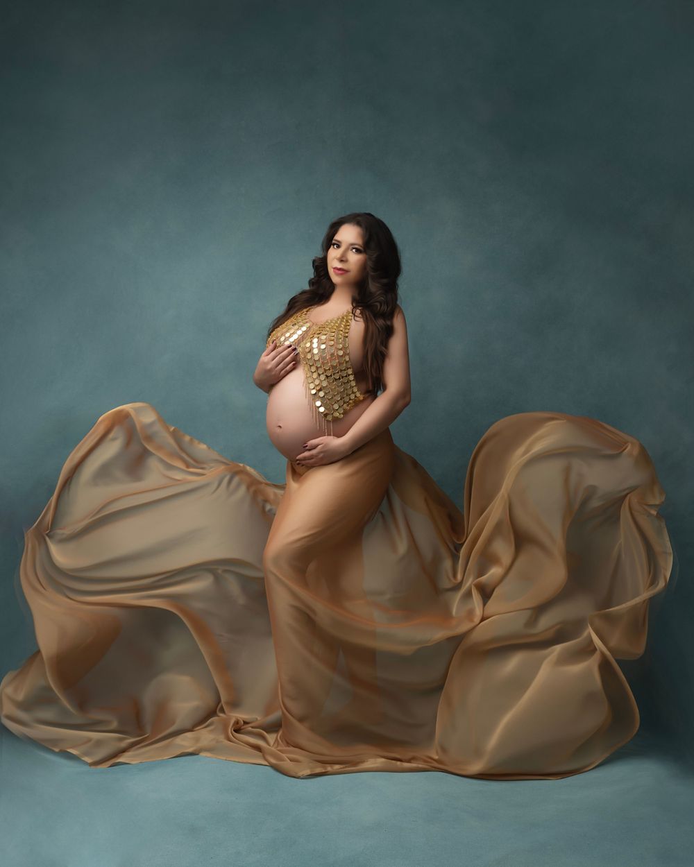 Artistic maternity portrait with billowing gold fabric creating dramatic waves against teal background.