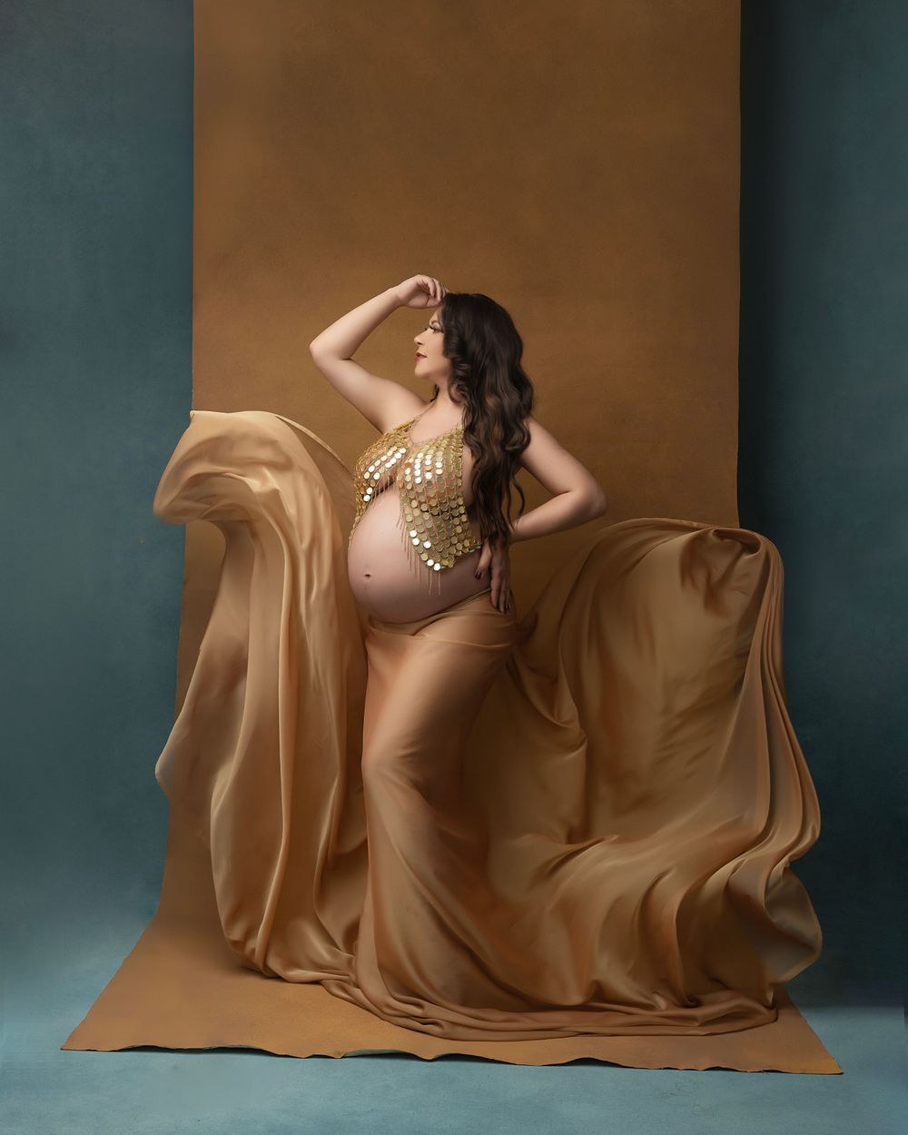 Reclining maternity pose with golden fabric draping gracefully against dark teal studio background.
