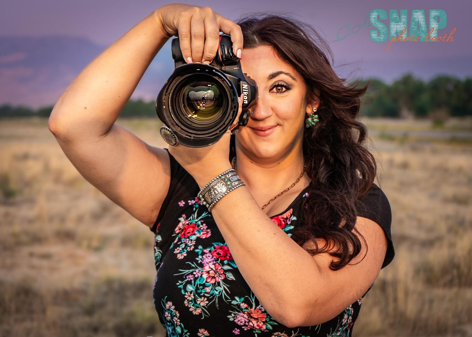 Professional photographer with camera and floral dress poses outdoors at sunset.