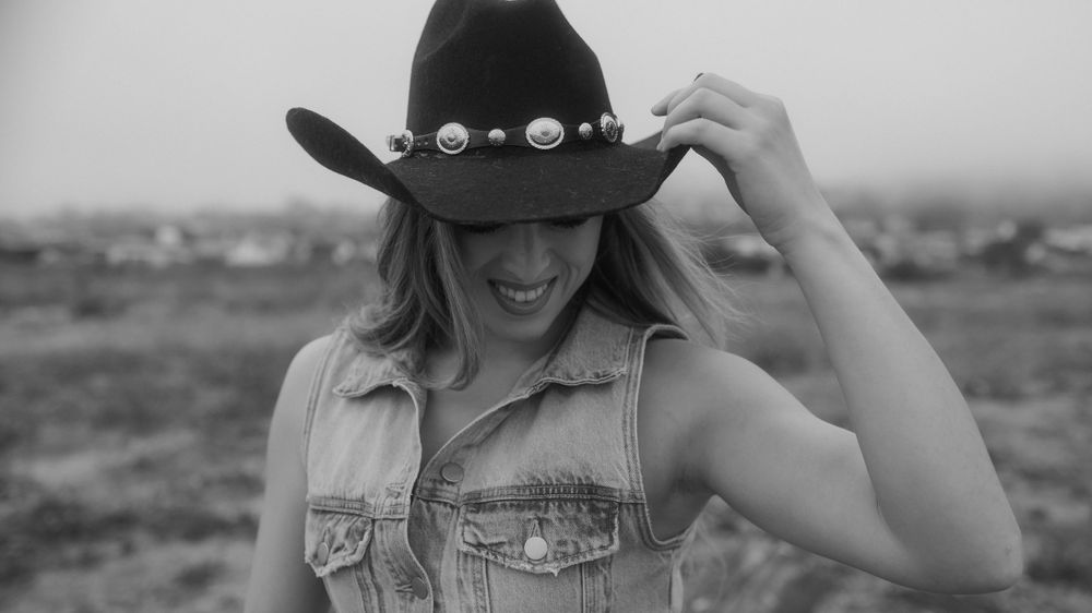 Black and white portrait in cowboy hat and denim vest showing joyful expression.