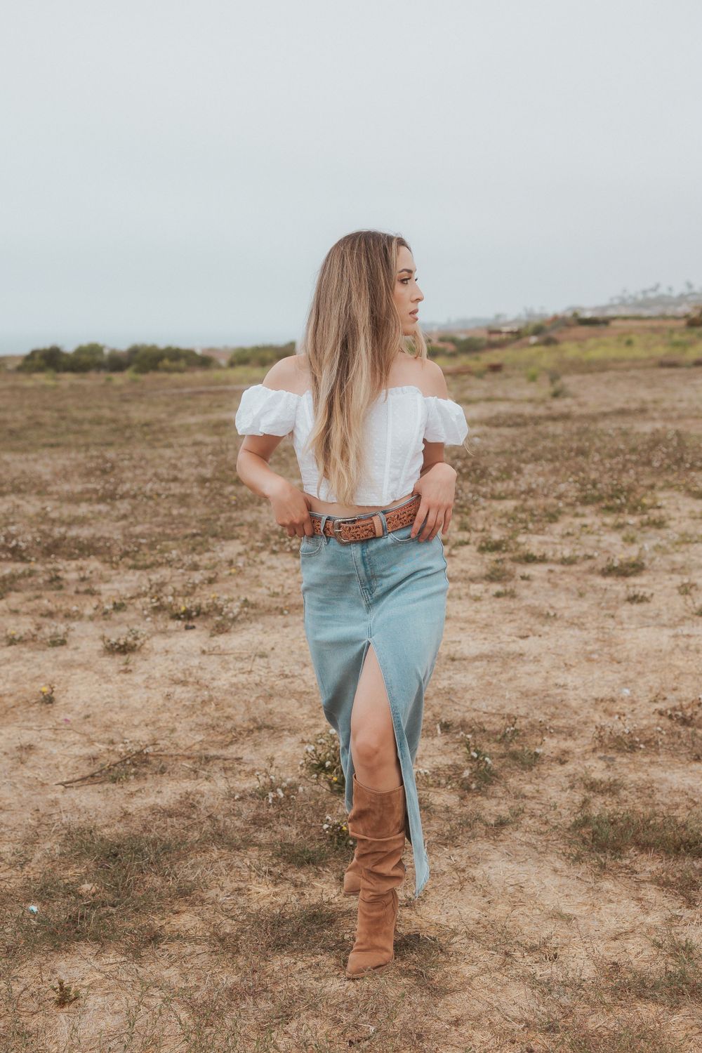 Country chic outfit with off-shoulder top, denim skirt and brown boots in desert setting.