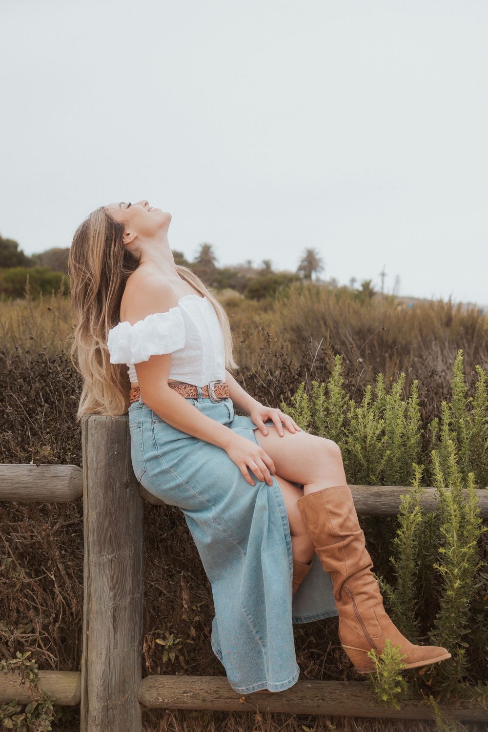 Rustic portrait on wooden fence wearing off-shoulder dress and brown cowboy boots.
