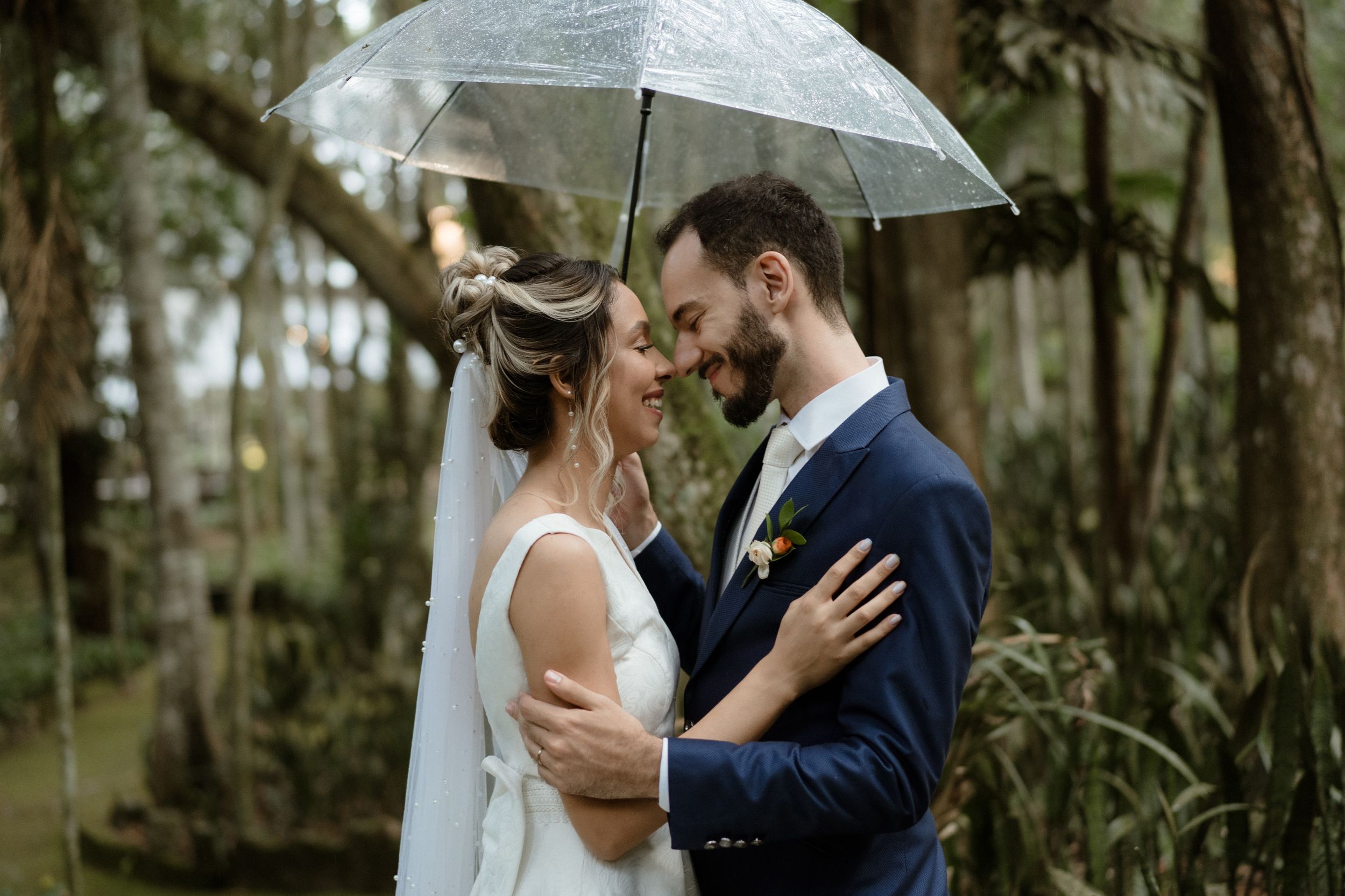 A couple shares an intimate moment under a clear umbrella in a lush forest setting during their wedding.