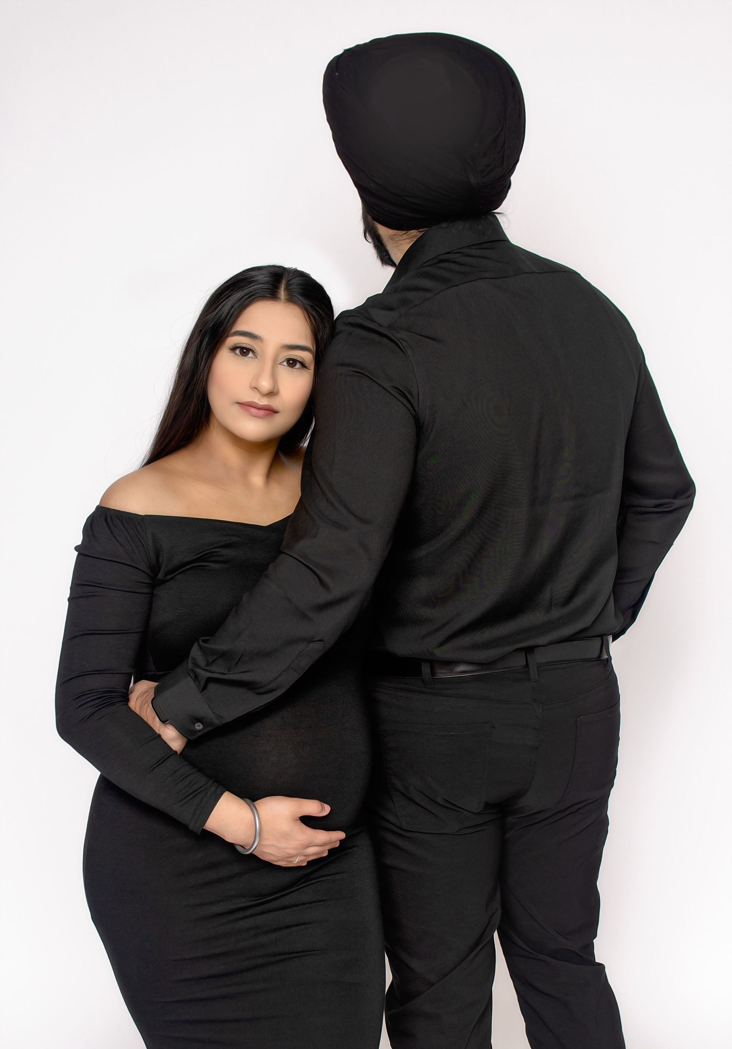 Couple in matching black outfits embracing against white background during maternity photoshoot.