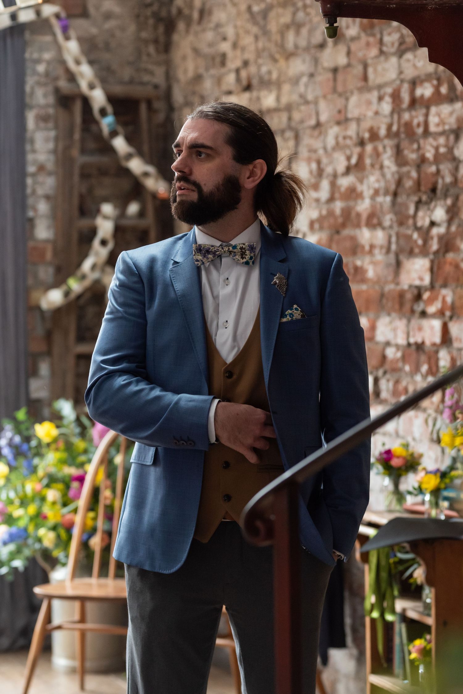 A person in a blue suit jacket and brown vest stands near floral arrangements at a wedding venue.