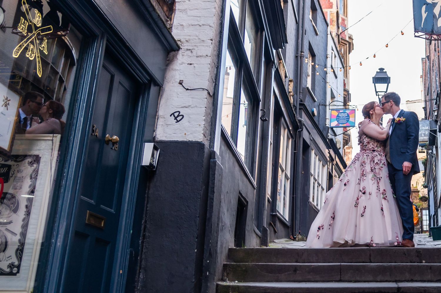A couple in wedding attire strolls through narrow city streets with blue-painted buildings.