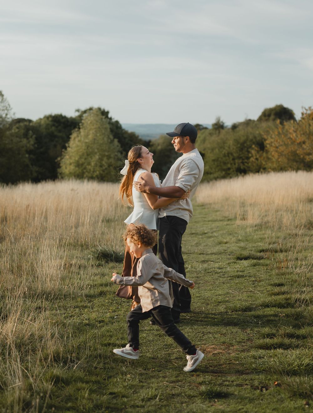 A couple embraces and spins joyfully in a sunlit meadow with tall grass and trees in the background.