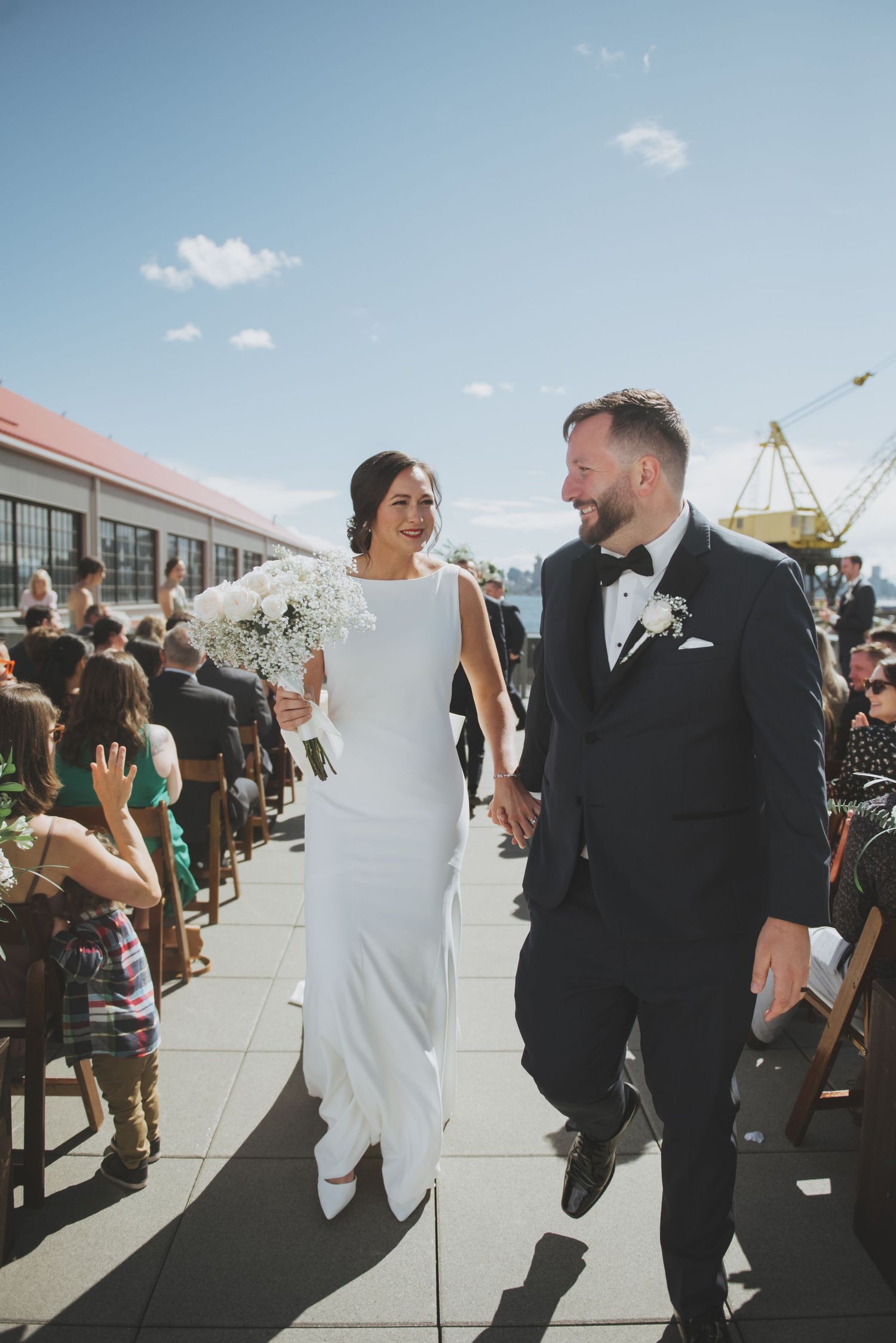 Newlyweds walking down aisle together with cityscape and industrial cranes in background.