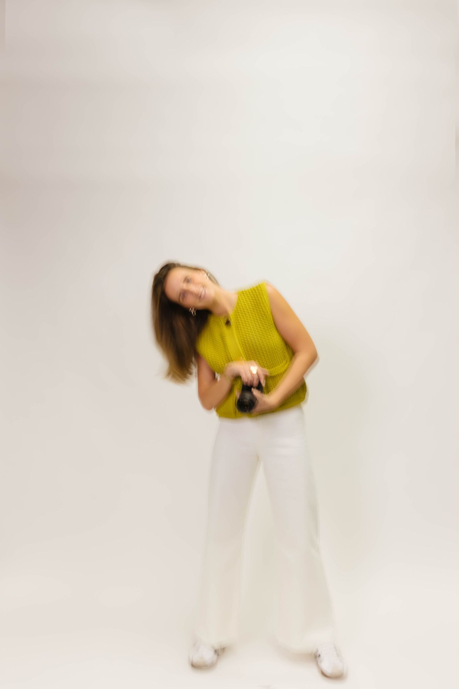 Motion-blurred photo of person in yellow top holding camera against white backdrop.