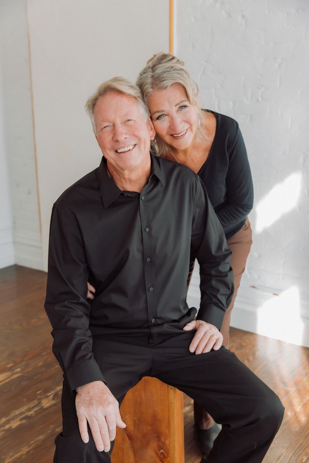A happy mature couple dressed in dark clothing pose together on a wooden stool in a bright sunlit room.