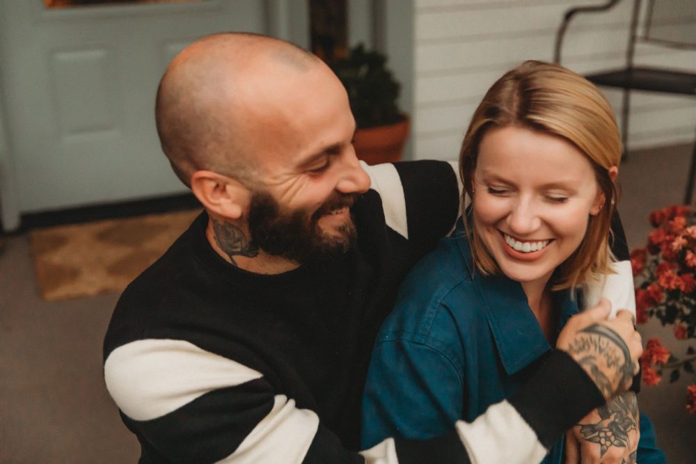 A couple sharing laughter while sitting together indoors wearing cozy winter sweaters.