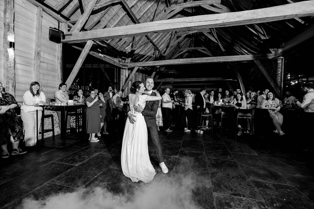 Black and white photo of a couple sharing their first dance on a foggy dance floor at a rustic barn wedding reception.