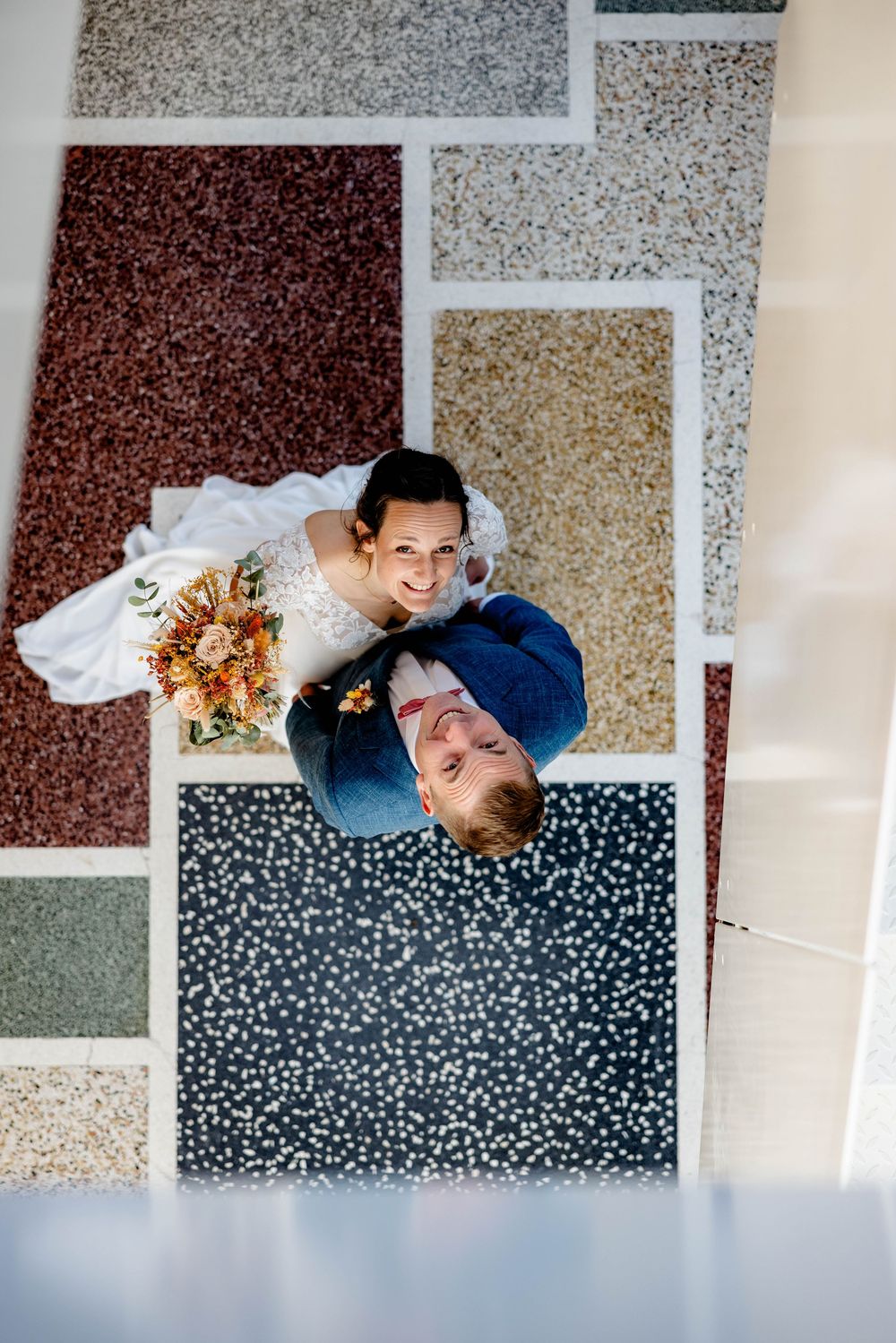 Aerial view of a smiling bride holding a bouquet and groom in blue suit standing on colorful terrazzo floor tiles.