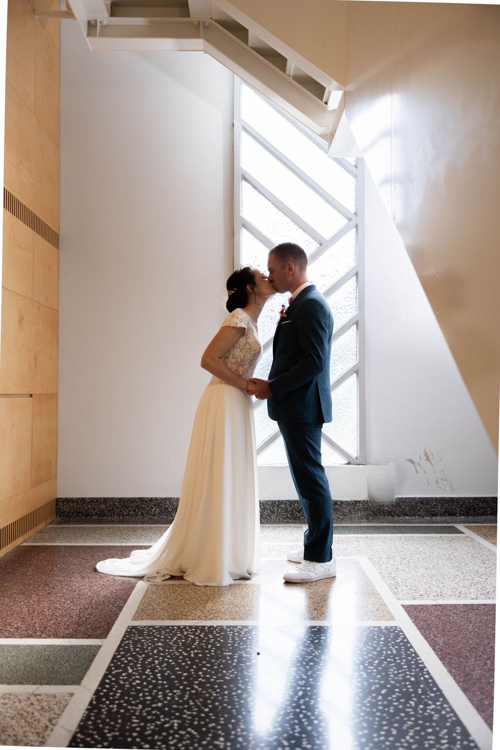 Bride and groom share a kiss beneath a modern staircase with geometric windows in soft natural light.