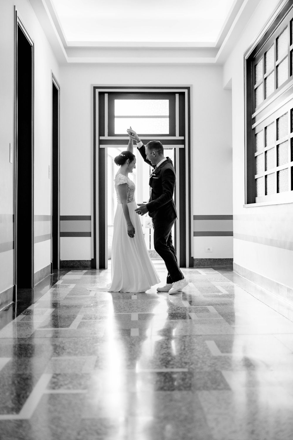 Black and white photo of bride and groom dancing elegantly in a sunlit hallway with marble floors.