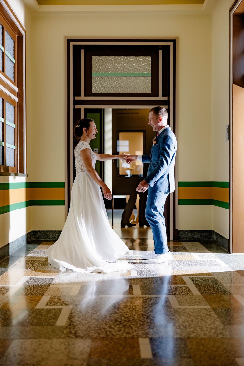 Bride in white gown and groom in blue suit share a tender moment in an elegant hall with marble floors.