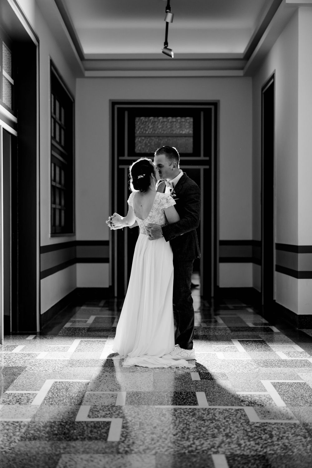 Black and white photo of bride and groom embracing in elegant hallway with patterned marble floor.
