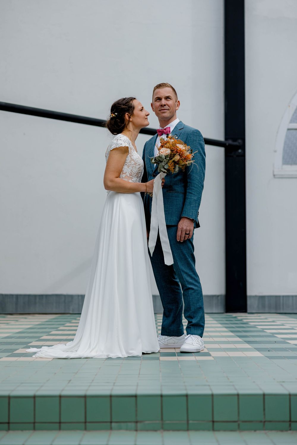 Bride in white gown holding bouquet stands with groom in blue suit and sneakers on tiled floor.