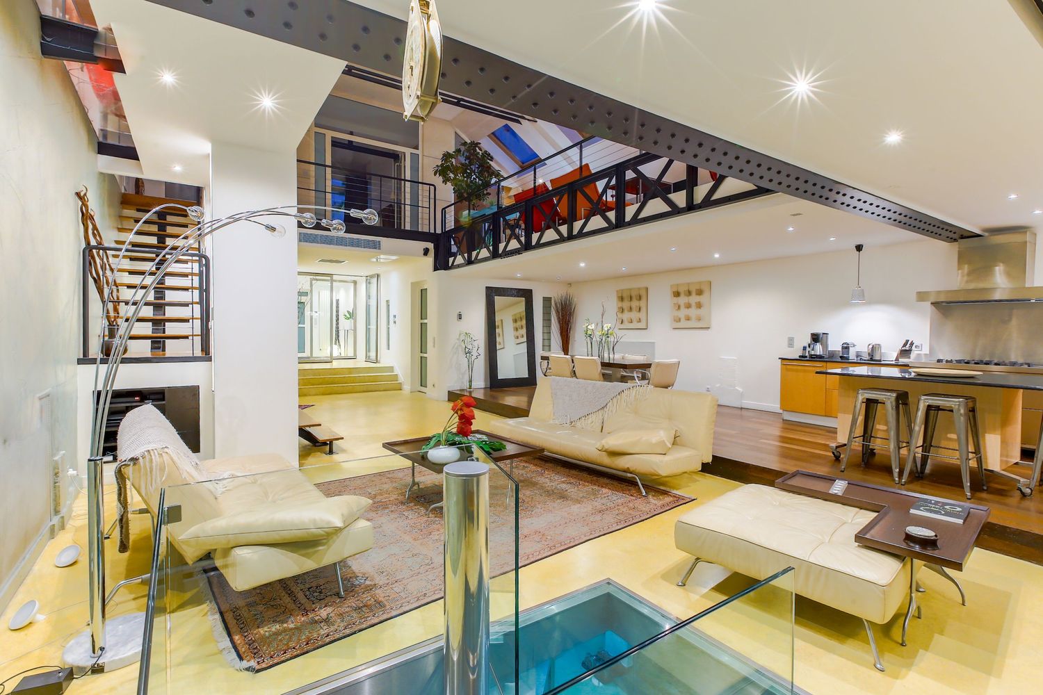 Luxury loft interior featuring glass floor panel, white sofas, mezzanine, and open-plan kitchen area.