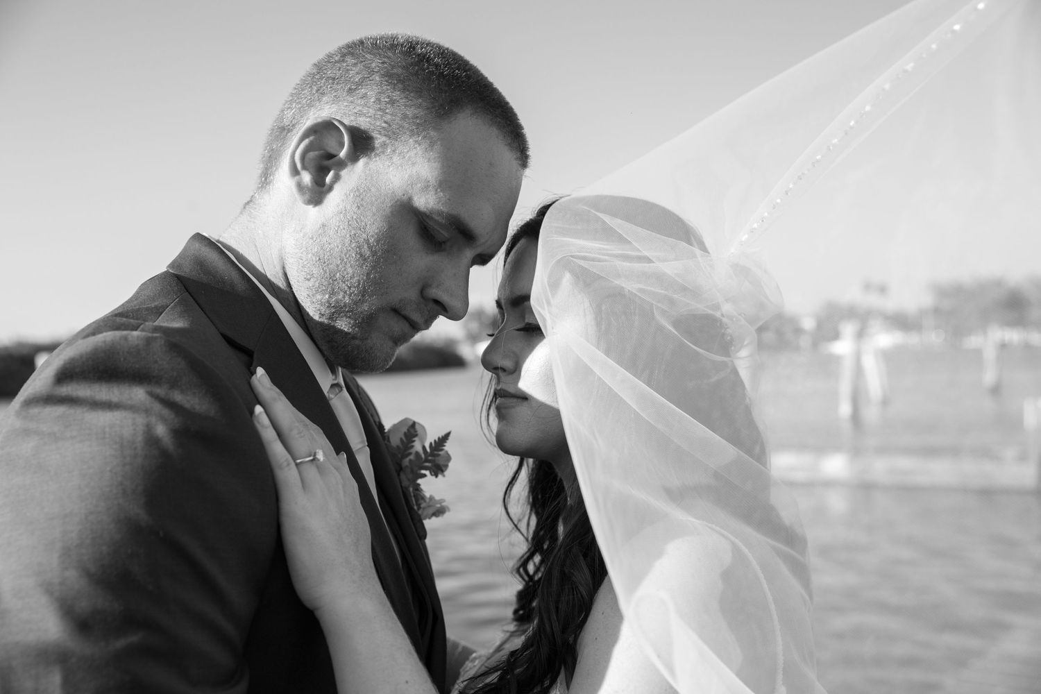 A black and white intimate wedding portrait with a flowing veil against a waterfront backdrop.