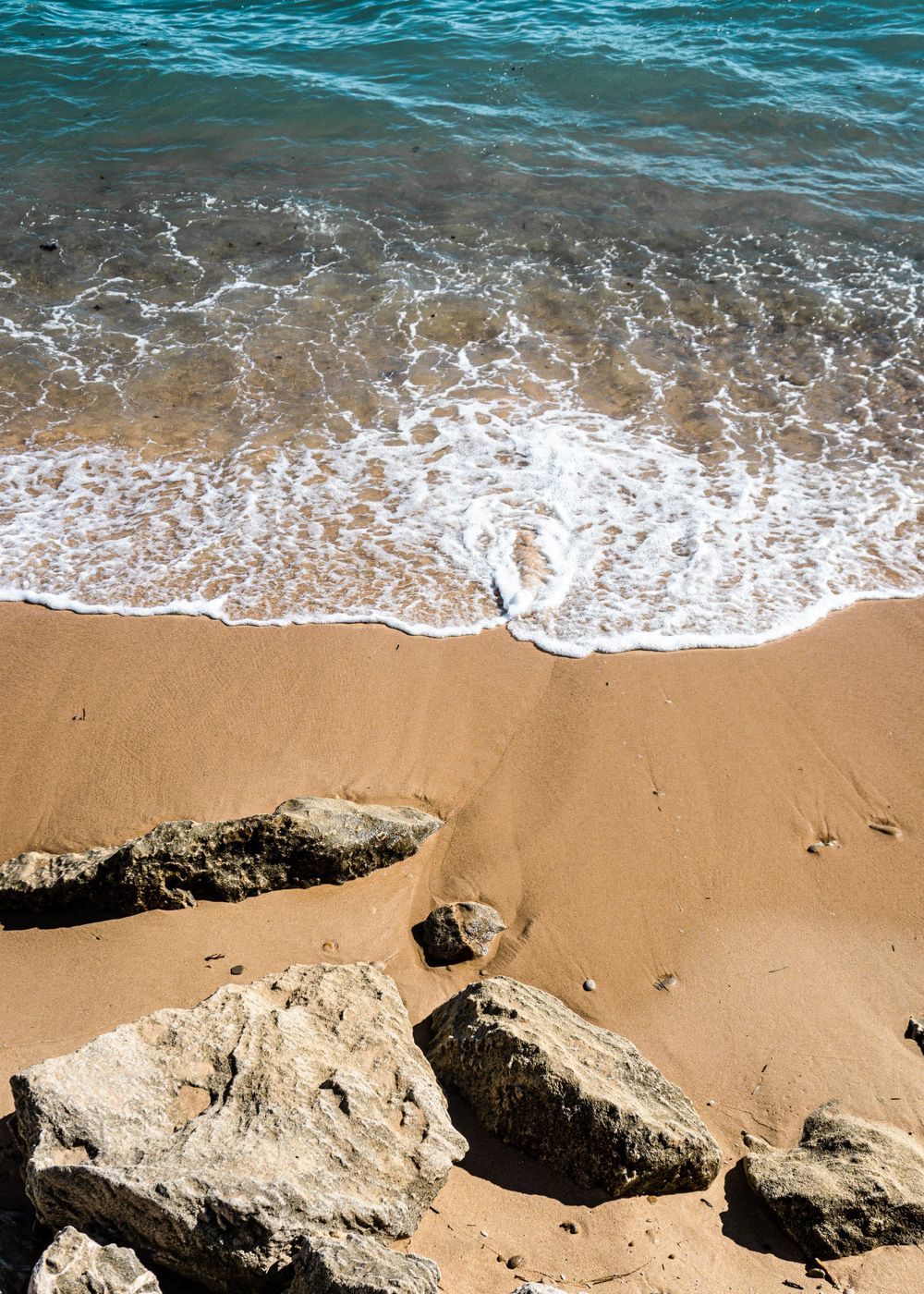 Turquoise waves washing up on sandy beach with large rocks in foreground.