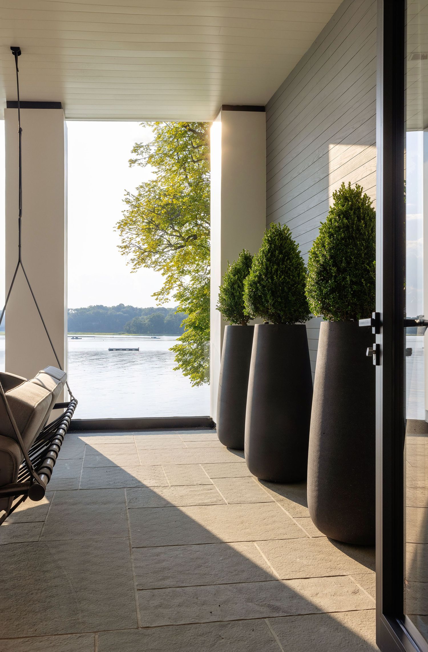 A modern lakeside porch with tall dark planters holding cone-shaped topiaries and a hanging chair. Sarza Rye NY