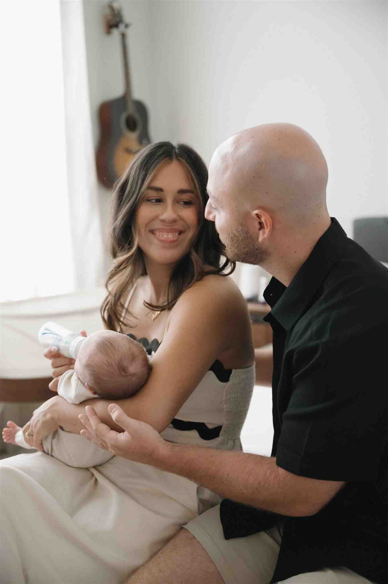A couple shares a tender moment with their newborn baby in a bright room with a guitar mounted on the wall.