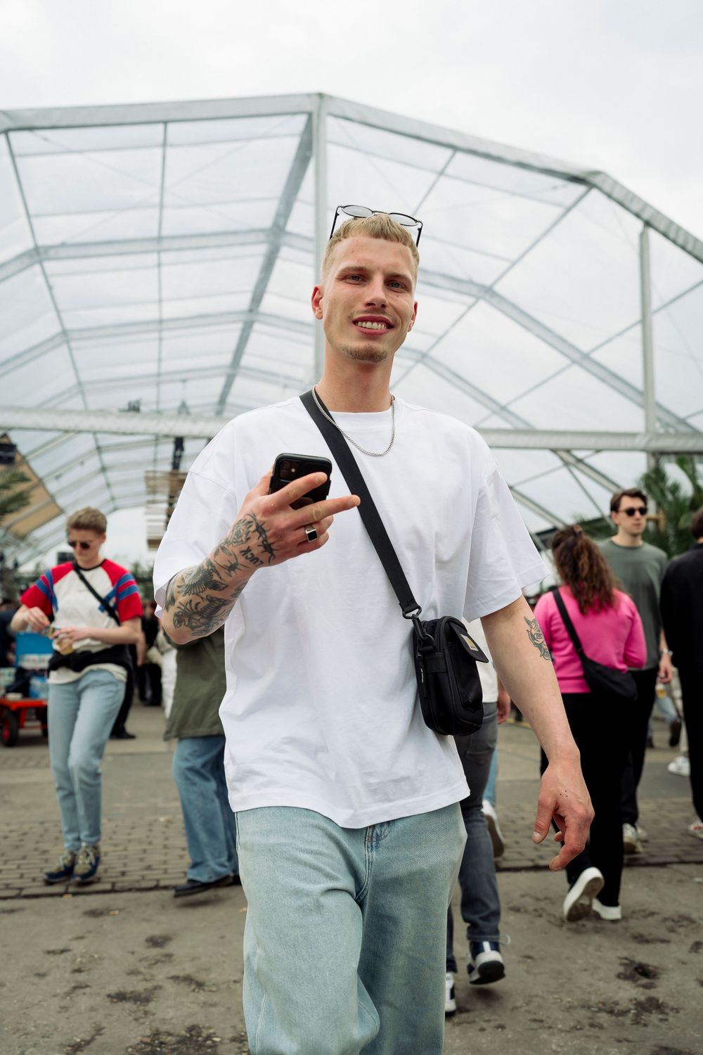 Person in white t-shirt and light pants standing under transparent tent structure with crowd in background.