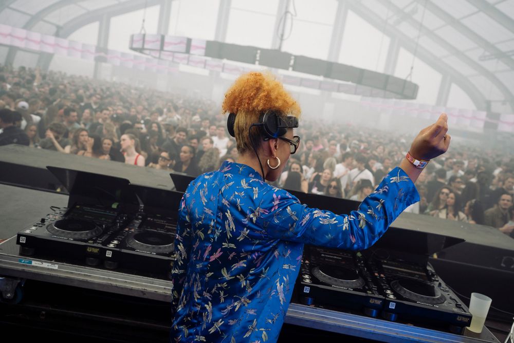 DJ wearing vibrant blue jacket raises arms while performing.
