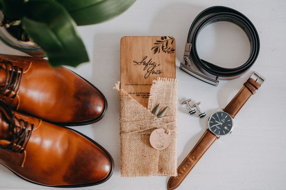 Brown leather dress shoes, wristwatch, belt and wooden accessory holder arranged on white surface.